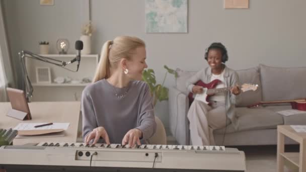 中程度の遅いの若いです白人女性演奏キーボードと彼女のアフリカ系アメリカ人のガールフレンド演奏電気ギター 練習曲でホーム音楽スタジオ — ストック動画