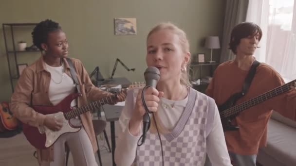 中等速度的多民族乐队三个人在室内摄像机前表演 年轻的白人妇女用麦克风唱歌 而她的朋友用电吉他和低音吉他手 — 图库视频影像