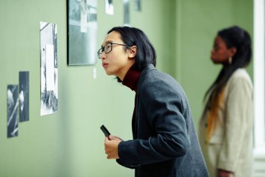 Gözlüklü yakışıklı genç Asyalı adamın portresi sanat galerisini ziyaret ediyor ve duvardaki siyah beyaz fotoğrafa odaklanıyor.