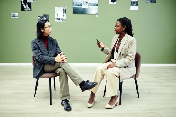 アフリカ系アメリカ人の女性が椅子に座ってボイスレコーダーを持っていて 現代の若いアジア人写真家にインタビューしています — ストック写真