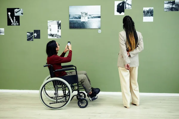 Unrecognizable young Asian man with disability and stylish Black woman visiting contemporary photography exhibition in art gallery looking at photos and scanning qr code