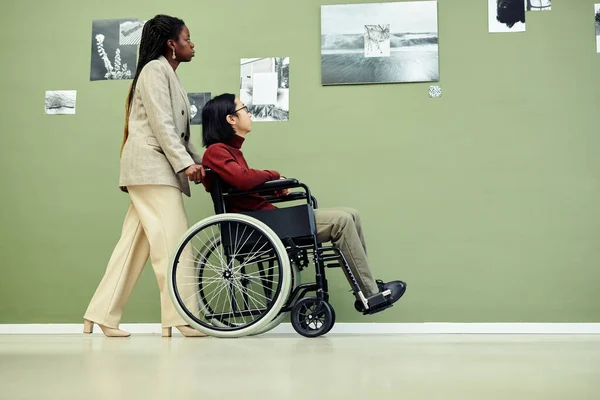 Side view of young African American woman and her Asian male friend with disability in wheelchair visiting modern black and white photography exhibition