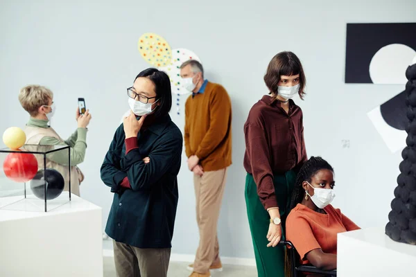 Group of diverse multi-ethnic people wearing protective masks examining various contemporary art objects at exhibition in museum