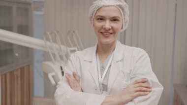 Beyaz laboratuvar önlüğü giyen, gülümseyen beyaz beyaz dişçi ve modern diş muayenehanesinde elleri katlanmış kameraya poz veren tek kullanımlık tıbbi şapkalı kadın portresi.