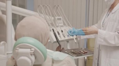 Tek kullanımlık tıbbi başlık ve yüz maskesi takan, eldivenlerini çıkaran, modern diş kliniğinde oturan hasta için diş muayenesini bitiren orta yaşlı bir dişçi.