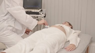 Kadın jinekolog yavaş yavaş ultrason taraması yapıyor ve modern klinikteki tıbbi kanepede yatan hamile bir kadına ultrason taraması yaparken monitöre bakıyor.