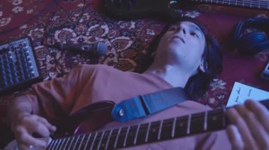 Yukarıdan gelen, uzun kahverengi saçlı, elektro gitar çalan, Neon Mor ışıklı, antika bir halı üzerinde uzanan hippi genç adam.