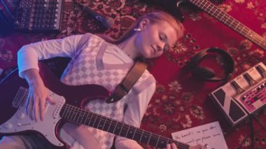 Yukarıdan gelen y2k 'nin orta yavaşlığında genç bir kadın elektro gitar çalarken gülümserken her yerde aşk şarkısı sözleri olan kırmızı halıda yatıyor.