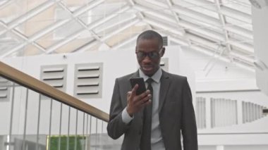 Modern iş merkezinde yürürken cep telefonuyla konuşan resmi giyimli ve gözlüklü genç Afrikalı Amerikalıların orta derecede yavaşlaması.