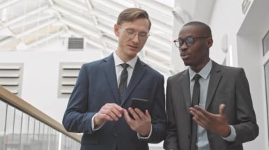 Modern iş merkezinde yürürken resmi giyinmiş iki zarif iş adamının orta yol yavaşlığı. İş adamlarından biri de akıllı telefonunu kullanıyor.