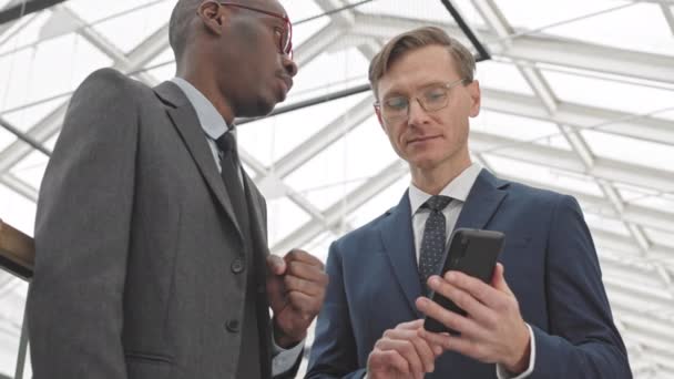 两个成功的青年商人站在现代商务中心 一边看着智能手机 一边慢吞吞地谈着话 — 图库视频影像