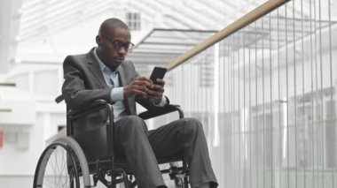 Engelli genç Afrikalı Amerikalı iş adamının yavaş yavaş resmi kıyafetleri ile çağdaş iş merkezinde tekerlekli sandalyede otururken telefon görüşmesi yapıyor.