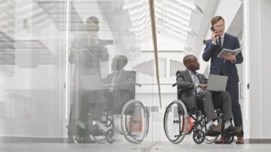 Tekerlekli sandalyedeki Afrikalı Amerikalı işadamı ve Kafkasyalı iş ortağının çağdaş iş merkezinde proje stratejisini tartışırken dizüstü bilgisayarını kullanması.