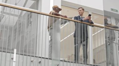 Afrikalı Amerikalı teslimatçı, kendinden emin beyaz iş adamlarına iş belgelerini teslim ederken, onlar için imza dağıtırken, çağdaş iş merkezinde cam korkulukların yanında dikilirken.