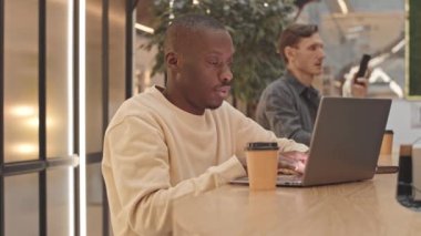 Yan görünüş, kafede dizüstü bilgisayarla çalışırken günlük giysiler içinde kahve içen Afrikalı Amerikalı erkek serbest çalışanın belini yavaş yavaş kaldırır.