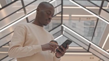 Düşük açılı, ağır ağır, kapüşonlu, akıllı telefonlu, kahve içen ve camdan tavanlı lüks iş merkezine bakan siyah bir adamın portresi.