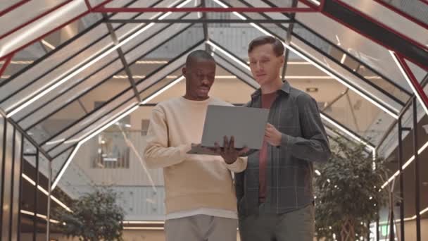 两个不同的男性设计师在使用笔记本电脑的同时 在玻璃三角天花板的现代商务中心完成共同的项目 其速度为中等 — 图库视频影像