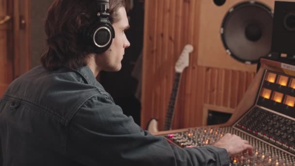 サイドビューウエストアップヘッドフォンで音楽プロデューサーのスローモーションミキシングコンソールを使用しながら彼の頭をうなずき スタジオで新しいトラックを録音 — ストック動画
