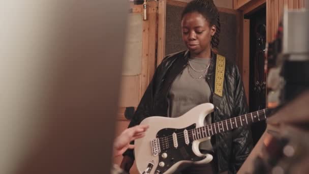 穿着皮夹克的非洲裔美国女吉他手在专业音乐录音室录制新歌的同时 调整电吉他 并与音乐制作人交谈 — 图库视频影像
