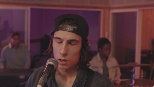 マイクで歌っている若い感情的なロックミュージシャンの減速を傾け ベースギターを演奏し 音楽スタジオで彼のバンドと一緒に演奏する — ストック動画