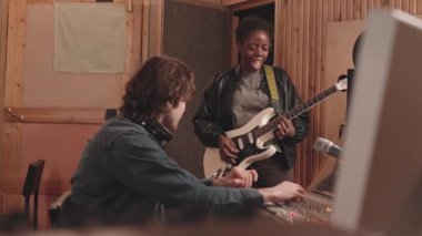 Müzik kayıt stüdyosunda elektro gitar çalan genç siyahi kadının orta yavaşlığı ses mühendisi karıştırma konsolunu çalıştırırken