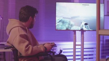 Tekerlekli sandalyedeki Kafkas oyuncusunun karanlık oyun salonunda neon ışıklı fps oyunu oynarken kumanda kullanmasının yavaşlığı