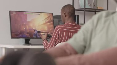 Meslektaşı ön planda bulanık video oyunları geliştirirken, Afrikalı Amerikalı bir adamın PC 'de kumandayla fps oyunu oynaması orta görünüm yavaşlığı