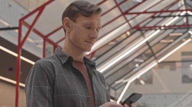 Günümüz iş merkezinde cam üçgen tavanlı, akıllı telefonlara göz atan, günlük giysili, genç beyaz adamın yavaşlığını kaldır.