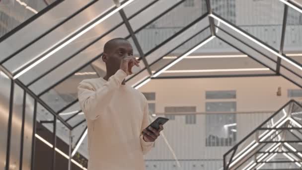 穿着运动衫的黑人年轻人站在现代商业中心 站在玻璃三角天花板的智能手机上一边喝着外卖杯咖啡 一边慢吞吞地喝着低角度的咖啡 — 图库视频影像