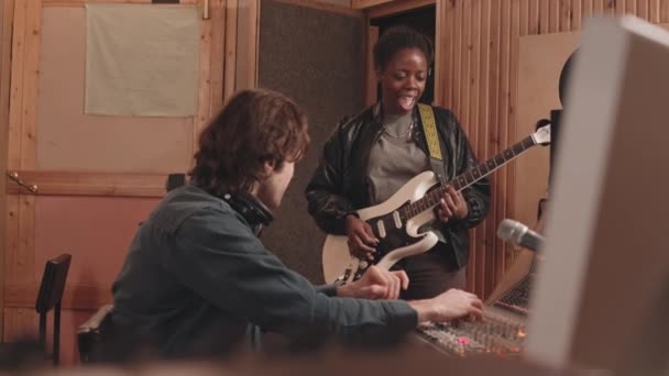 中等速度的年轻黑人妇女在音乐录音室弹奏电吉他 而音响工程师在操作混音控制台 — 图库视频影像