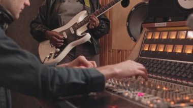 Profesyonel müzik stüdyosunda yeni parça kaydedilirken elektro gitar çalan ve müzik yapımcısının ses mikserini ayarlayan tanınamayan Afrikalı Amerikalı kadının yavaşlaması.