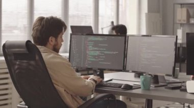 Bilgisayar oyunu geliştirirken ofiste iki bilgisayar monitörü önünde oturup klavyede program kodları yazan erkek programcının dikiz görüntüsü yavaşlaması