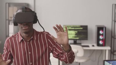 Modern programcılar ofisinde video oyunu oynarken VR kulaklıklı genç siyahi adamın orta derecede yavaşlaması.