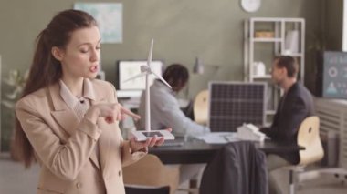 Elinde rüzgar türbini modeliyle genç beyaz kadın mühendisin orta yavaşlığında kameraya bakıp alternatif enerji kaynaklarından bahsederken meslektaşları arka planda iş birliği yapıyor.
