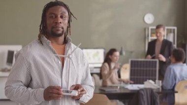 Afrika kökenli Amerikalı mühendis rüzgar türbini modeli ile kameraya alternatif enerji kaynakları hakkında konuşurken çeşitli meslektaşları arka planda çalışıyor.