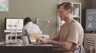 Yan görüş, ofisteki ekolojik proje üzerinde çalışırken dizüstü bilgisayarda yazan genç alternatif enerji mühendisinin yavaşlığını arttırıyor. Meslektaşı arka planda dünya haritasıyla masada oturuyor.