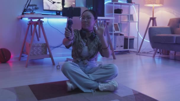 ビデオブログを記録しながら 加入者にゲームやサイバー空間について話している15歳のアジアの少女のスローモ 背景にコンピュータ画面上のFpsゲームと彼女の暗い部屋でラグに座って — ストック動画