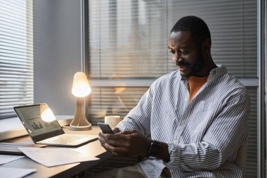 Ev işyerinde geç saatlere kadar çalışırken akıllı telefon kullanan gülümseyen siyah girişimcinin portresi lambayla aydınlatılıyor, kopyalama alanı