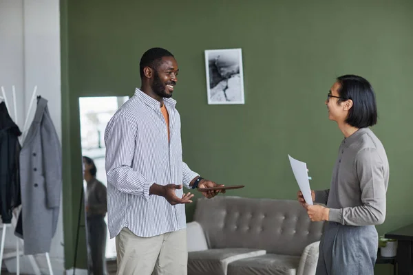 Waist up portrait of two ethnic business people discussing work and smiling while standing in modern office against green wall