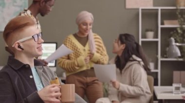İş belgelerini inceleyen ve kahve molası sırasında sohbet eden, modern ofiste birlikte çalışan genç yetişkin iş arkadaşlarından oluşan çok ırklı bir ekip.
