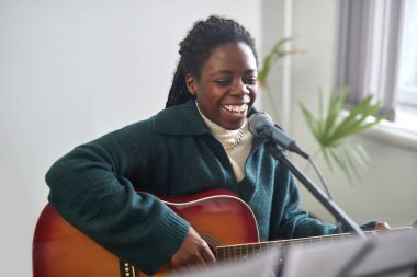 Afrikalı genç bir kadın mikrofona neşeli bir şarkı söylüyor ve sınıfta otururken gitar çalıyor.