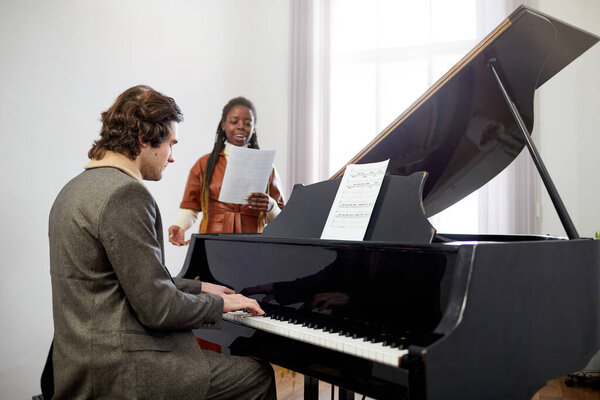 Учитель музыки играет на фортепиано, а его ученик поет песню, стоя за фортепиано во время урока музыки