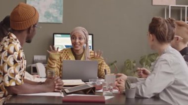 Toplantı sırasında ofis masasında oturup yeni projeyi tartışan orta yavaş yaratıcı takım. Başörtüsü takmış gülümseyen, başörtüsü takan, dizüstü bilgisayarlı genç bir kadın.