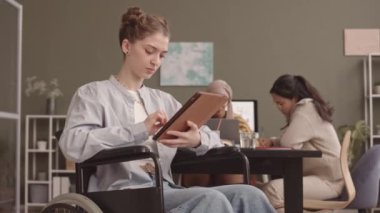 İş arkadaşları çalışma masasında arka planda çalışırken, tekerlekli sandalyede kameraya gülümseyen beyaz bir kadının orta yavaş portresi.