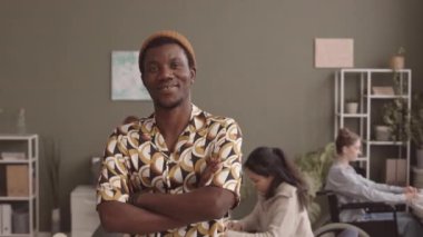 Hippi kıyafetli, turuncu şapkalı, modern ofiste çeşitli iş arkadaşlarıyla çalışırken kameraya poz veren neşeli siyahi bir adamın orta yavaş portresi.