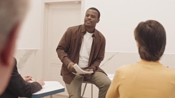 教室で数学のレッスンを受けている高齢の男性と女性のグループに話をしながら 彼の机にもたれているアフリカ系アメリカ人男性の遅い — ストック動画