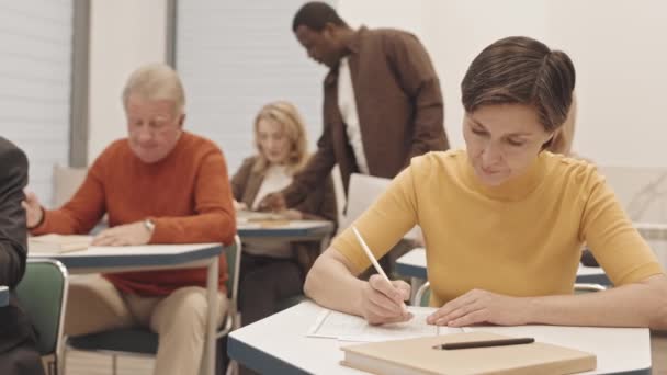 成熟した白人女性や他の大人の人々の腰をゆっくりと教室で数学のテストを行う机の上に1つずつ座っている 男性黒人教師机に沿って歩くとそれらを助ける — ストック動画