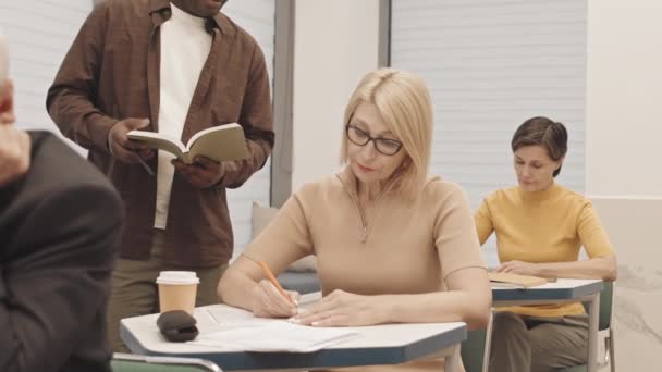 ウエストアップゆっくりのエレガントなブロンド成熟した女性で眼鏡をかけます数学テストしながら座っていますデスクで教室で他の大人とともに専門開発トレーニングコース — ストック動画