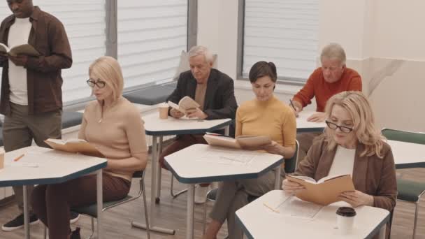 本や教材を持つ教室の机に一人ずつ座っている大人の男性と女性のグループの遅い上から トピックを説明する男性ブラック先生とレッスンを受けて — ストック動画