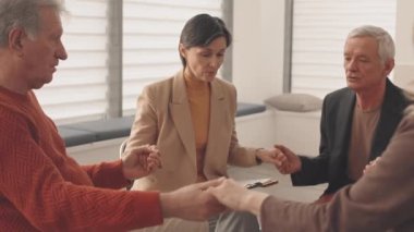 Profesyonel kadın terapist ve bir grup olgun insan destek grubu toplantısında el ele tutuşuyorlar.